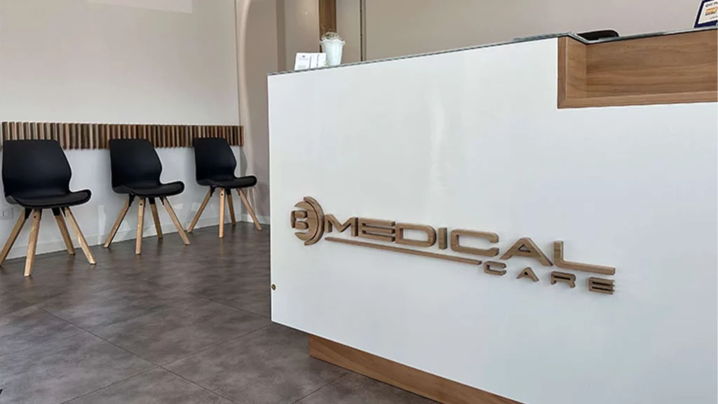 Logo B Medical Care tridimensionale con effetto legno, montato sul bancone bianco di una reception o studio medico, con sedie nell'area d'attesa, ideale per insegne luminose da interno.