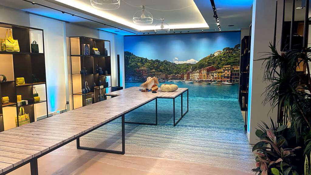 Interno di un negozio con espositori per borse e un grande murale che ritrae un paesaggio marino tipo Portofino, con pavimento stampato ad effetto acqua, ideale per insegne luminose da interno.