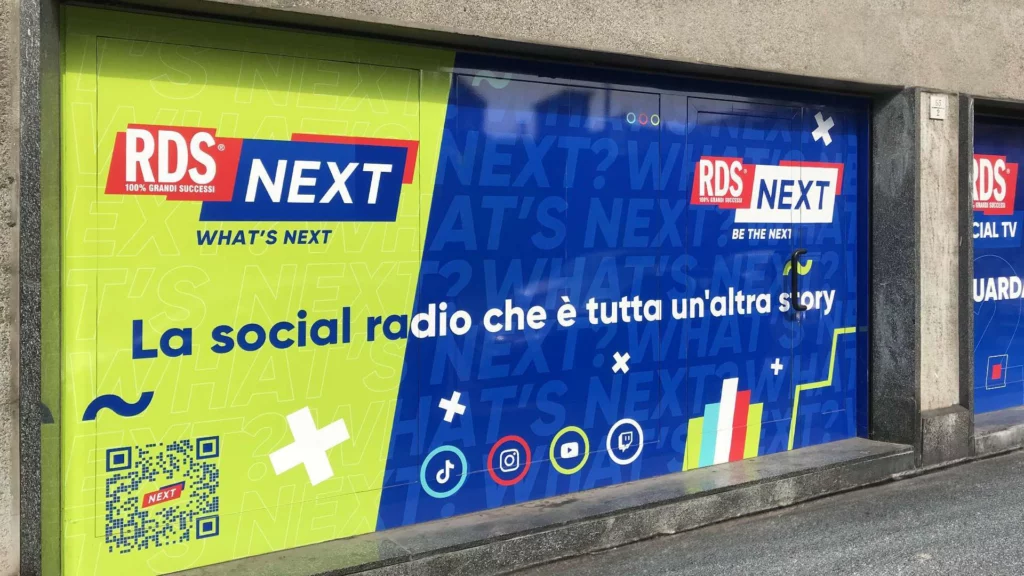 Serranda o porta di garage decorata con grafica adesiva RDS NEXT e slogan "La social radio che è tutta un'altra story", con QR code, ideale per cartelloni pubblicitari da esterno.
