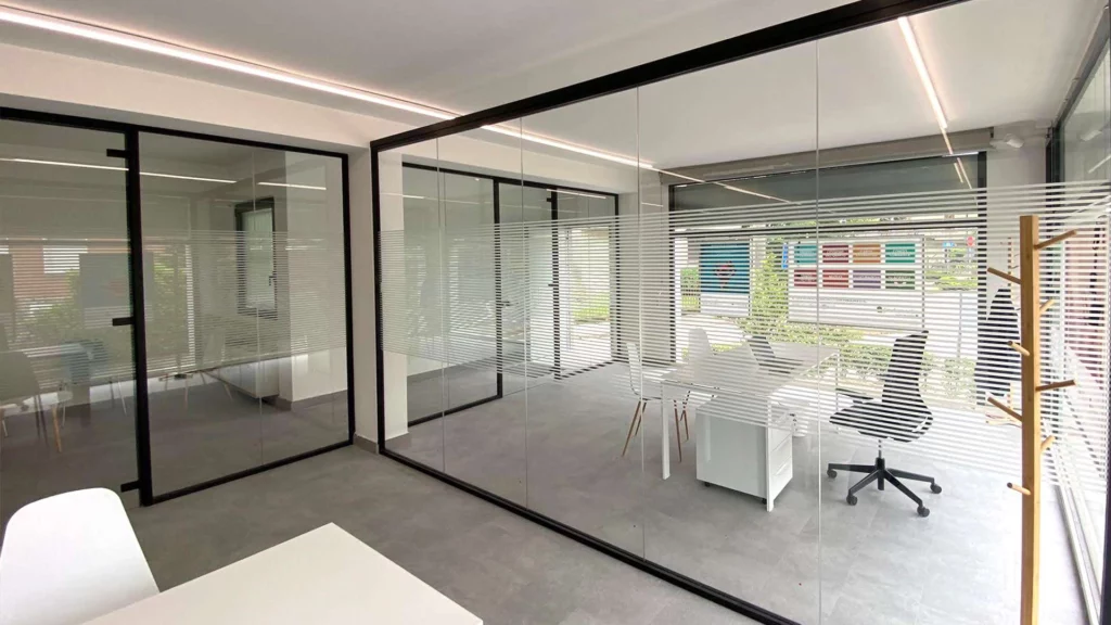 Ufficio moderno e luminoso con pareti divisorie in vetro e strisce orizzontali in pellicola adesiva, arredato con scrivania bianca e sedia da ufficio nera, ideale per insegne luminose da interno.