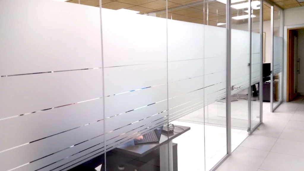 Corridoio d'ufficio con pareti divisorie in vetro decorate con pellicola adesiva smerigliata in strisce orizzontali trasparenti, con un laptop su un tavolino, ideale per insegne luminose da interno.