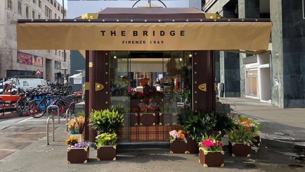 Chiosco o punto vendita The Bridge Firenze 1969 decorato in stile valigia di pelle marrone con tenda esterna, circondato da piante in vaso, ideale per insegne luminose da esterno.
