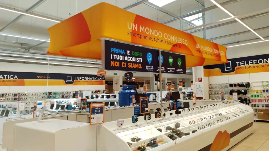 Struttura espositiva aerea luminosa nel reparto tecnologia di Unieuro, con la scritta UN MONDO CONNESSO e comunicazione sui servizi post-acquisto, ideale per insegne luminose da interno