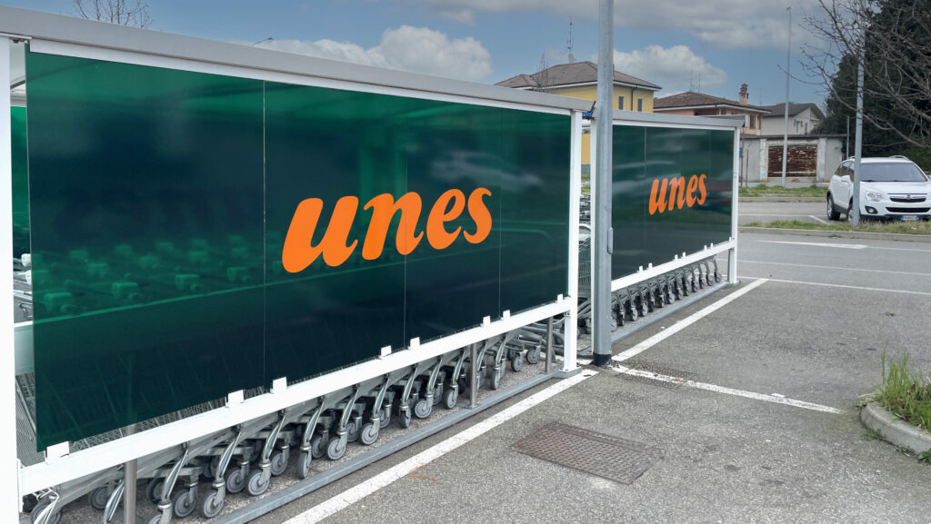 Telo pubblicitario verde con logo Unes arancione che copre un deposito di carrelli del supermercato ideale per decorazione auto