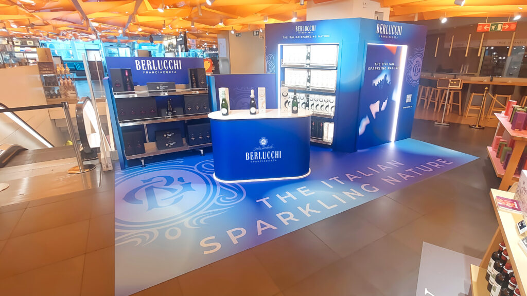 Stand espositivo Berlucchi Franciacorta con grafica blu luminosa e pavimento personalizzato con la scritta "The Italian Sparkling Nature" in un centro commerciale, ideale per insegne luminose da interno.